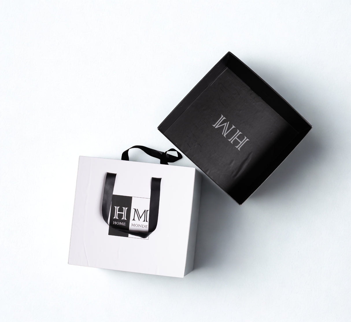A stylish white shopping bag with black handles and a HOME MONDE logo sits next to its matching box, both on a light background锟斤拷锟絧erfect for gifting the Premium Bedsheet & Cushion Covers Combo Set in its gift-read