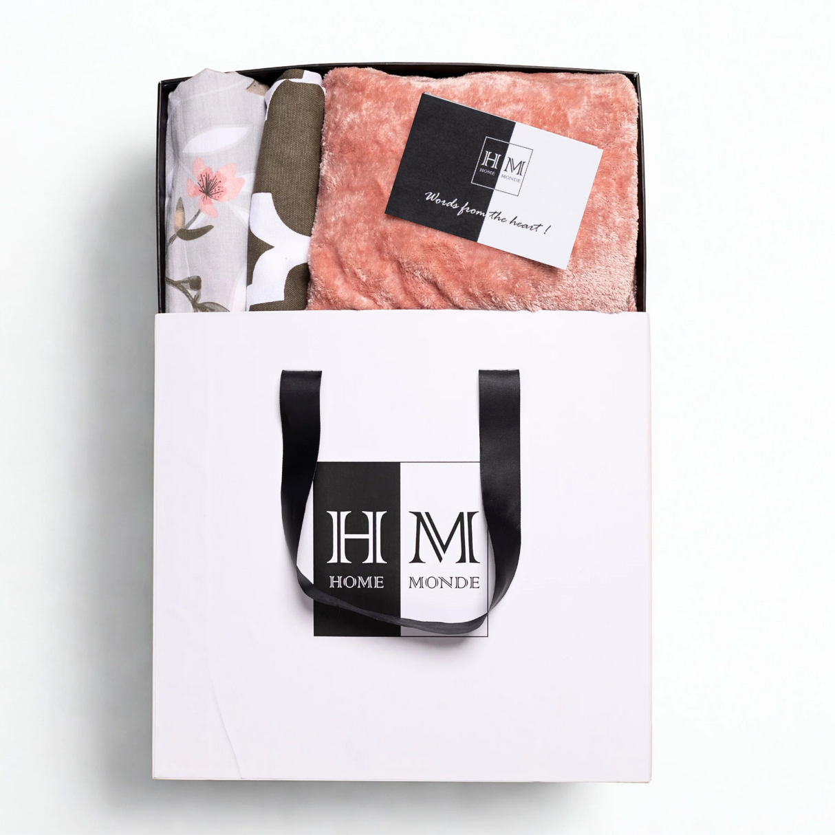A white gift bag with black handles and a HOME MONDE logo holds the Premium Bedsheet, Blanket & Cushion Cover Combo Set锟斤拷锟絝eaturing a pink fleece blanket, floral cotton bedsheet, and a 锟斤拷锟絎ords from the heart!锟矫