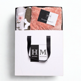 A white gift bag with black handles and a HOME MONDE logo holds the Premium Bedsheet, Blanket & Cushion Cover Combo Set锟斤拷锟絝eaturing a pink fleece blanket, floral cotton bedsheet, and a 锟斤拷锟絎ords from the heart!锟矫