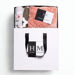 A white gift bag with black handles and a HOME MONDE logo holds the Premium Bedsheet, Blanket & Cushion Cover Combo Set锟斤拷锟絝eaturing a pink fleece blanket, floral cotton bedsheet, and a 锟斤拷锟絎ords from the heart!锟矫