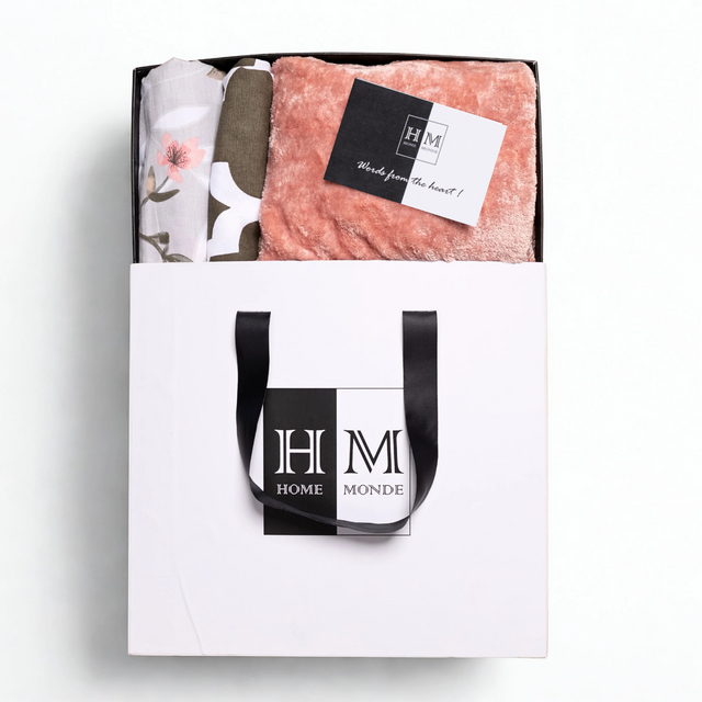 A white gift bag with black handles and a HOME MONDE logo holds the Premium Bedsheet, Blanket & Cushion Cover Combo Set锟斤拷锟絝eaturing a pink fleece blanket, floral cotton bedsheet, and a 锟斤拷锟絎ords from the heart!锟矫