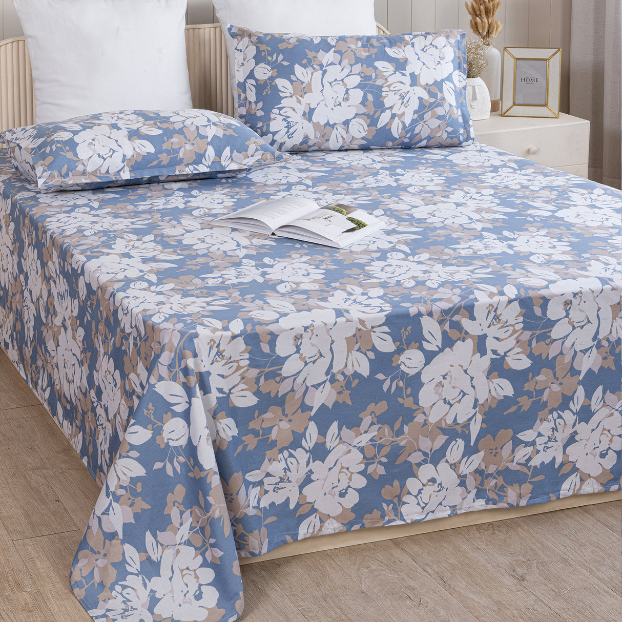 A neatly made bed features the Imprimer Blossom Haze - Bedsheet in blue and beige floral cotton with matching pillowcases, set in a cozy bedroom with wooden floors, a white nightstand, and an open book resting on the bed.
