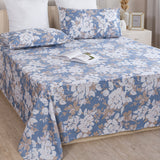 A neatly made bed features the Imprimer Blossom Haze - Bedsheet in blue and beige floral cotton with matching pillowcases, set in a cozy bedroom with wooden floors, a white nightstand, and an open book resting on the bed.