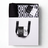 A white gift bag with black handles labeled HM Home Monde holds a folded rug, velvet cushion covers, and a "Made from the heart!" card锟斤拷锟絘 Premium Cushion Cover & Rug Combo Set in a gift-read