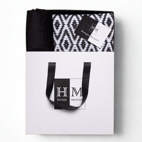 A white gift bag with black handles labeled HM Home Monde holds a folded rug, velvet cushion covers, and a "Made from the heart!" card锟斤拷锟絘 Premium Cushion Cover & Rug Combo Set in a gift-read