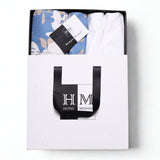 A white HOME MONDE box with black handles holds the Premium Bedsheet & Cushion Covers Combo Set锟斤拷锟絘 blue floral cotton bedsheet and branded tag锟斤拷锟絧hotographed from above on a white background, gift-ready for a