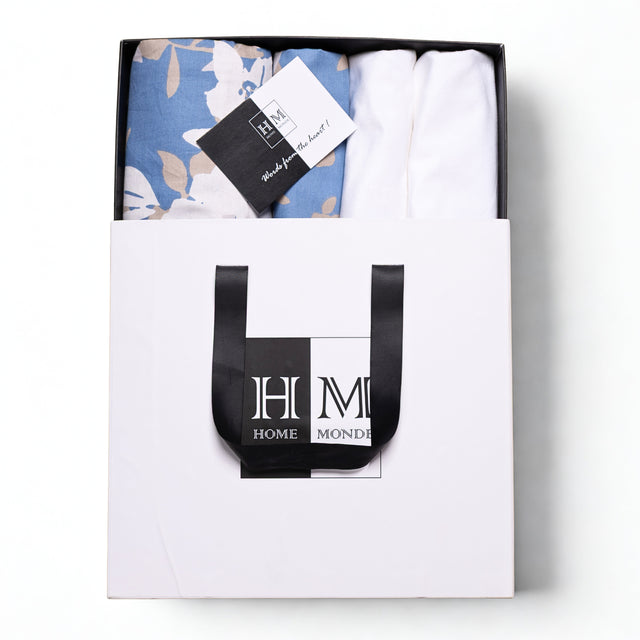 A white HOME MONDE box with black handles holds the Premium Bedsheet & Cushion Covers Combo Set锟斤拷锟絘 blue floral cotton bedsheet and branded tag锟斤拷锟絧hotographed from above on a white background, gift-ready for a