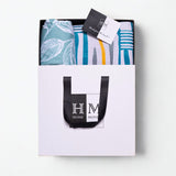 A white gift bag with black handles, containing a Premium Bedsheet & Runner Rug Combo Set in blue, teal, and yellow hues and a card on top. Gift-ready in a HOME MONI logo bag, it's the perfect present.