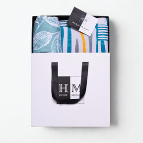 A white gift bag with black handles, containing a Premium Bedsheet & Runner Rug Combo Set in blue, teal, and yellow hues and a card on top. Gift-ready in a HOME MONI logo bag, it's the perfect present.