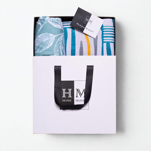 A white gift bag with black handles, containing a Premium Bedsheet & Runner Rug Combo Set in blue, teal, and yellow hues and a card on top. Gift-ready in a HOME MONI logo bag, it's the perfect present.